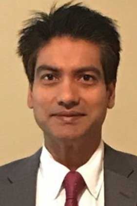 Dr. Mishra is a man of South Asian descent, wearing a gray suit and necktie.