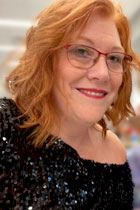 Tish is a Caucasian woman with red hair and pink-framed glasses. She is wearing a sparkling black blouse and smiling.