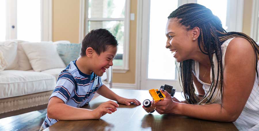 A child and caregiver play with trucks at a low table. Both are laughing.