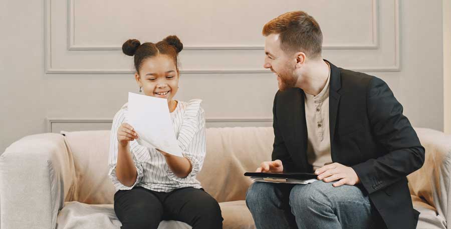 A Black American tween girl and a Caucasian male therapist sit together on a sofa, looking at a paper. They are smiling and laughing together.