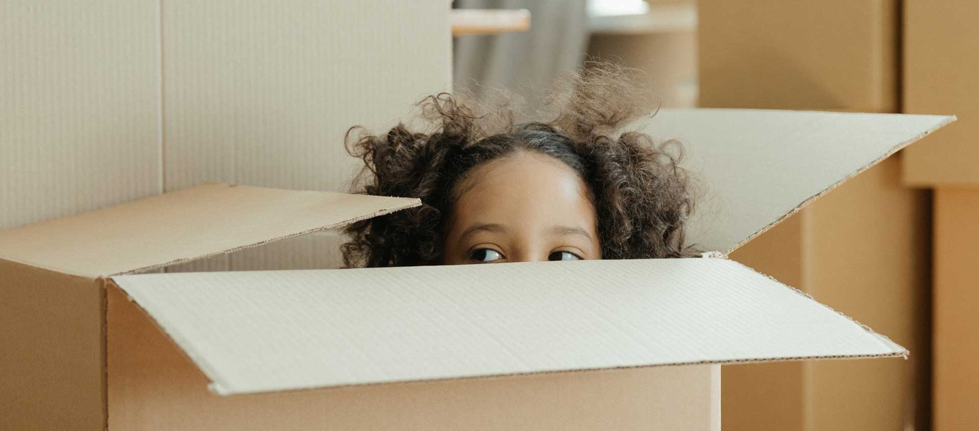 A girl with curly dark hair peeks over the edge of a moving box.