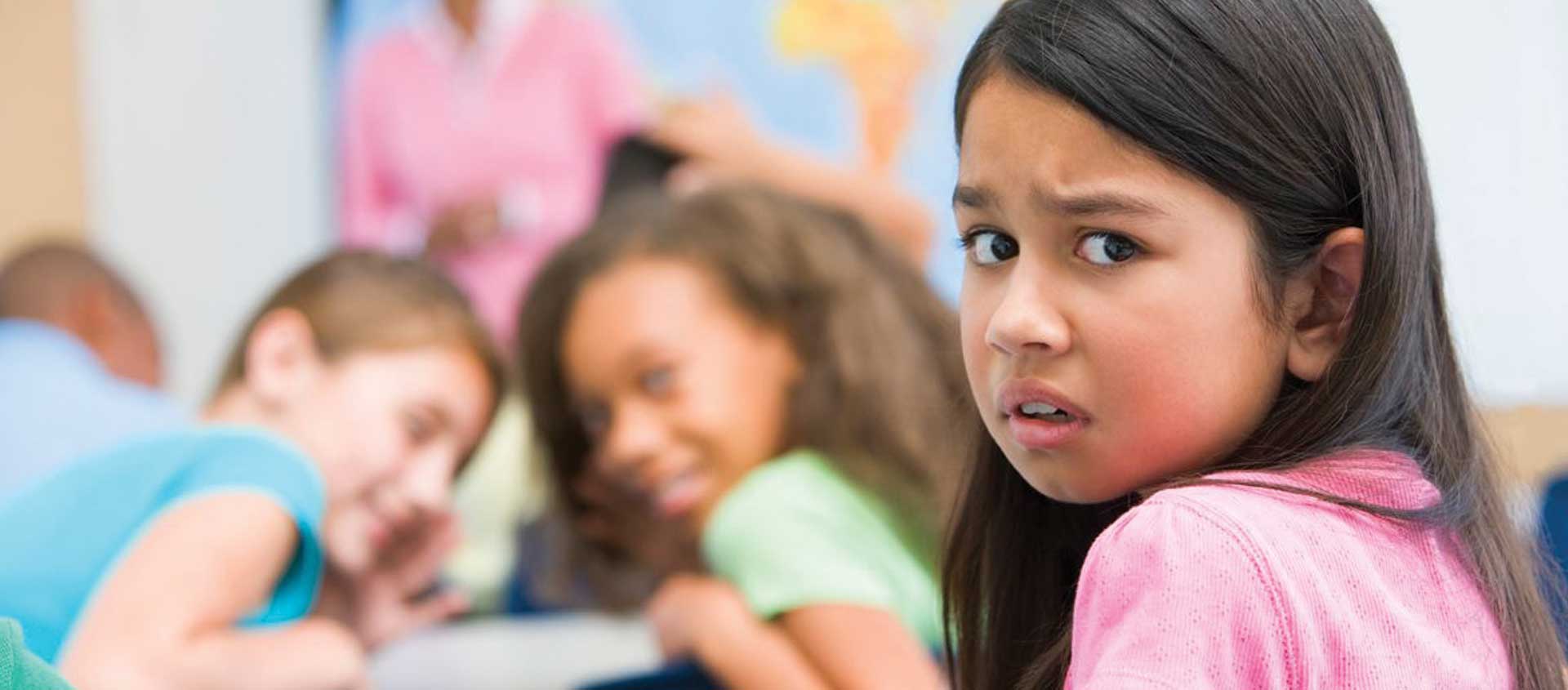 An elementary age girl looks upset while two classmates whisper behind her.