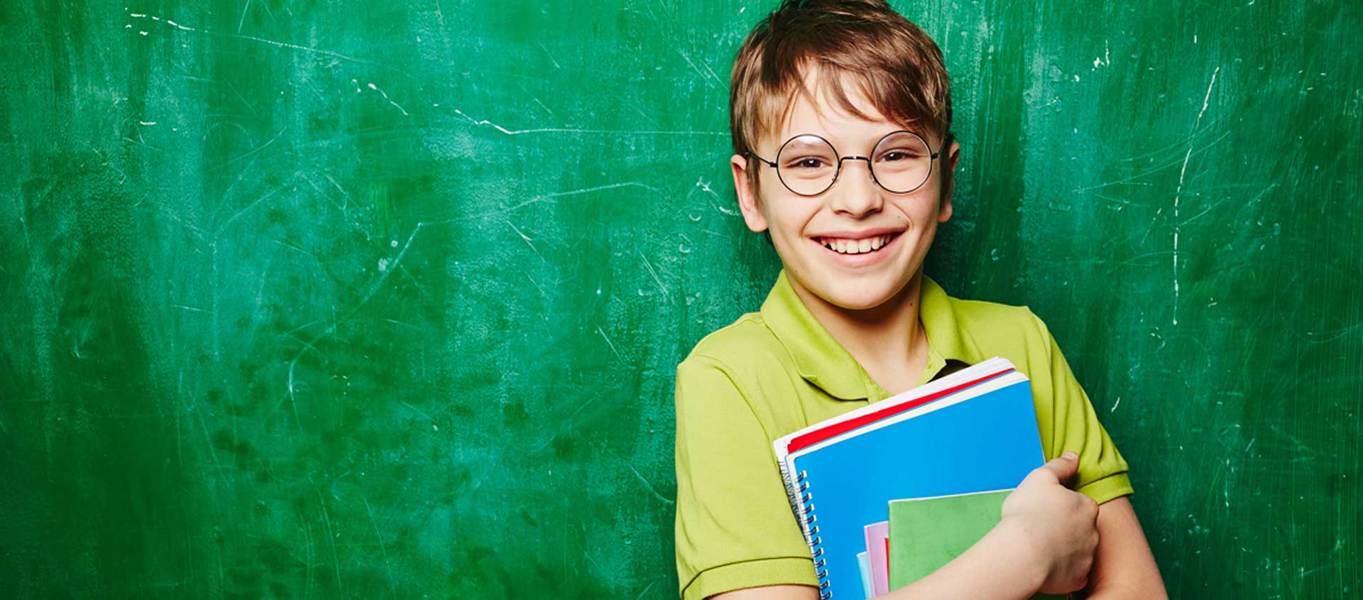 A Caucasian boy with glasses stands in front of a chalkboard, smiling