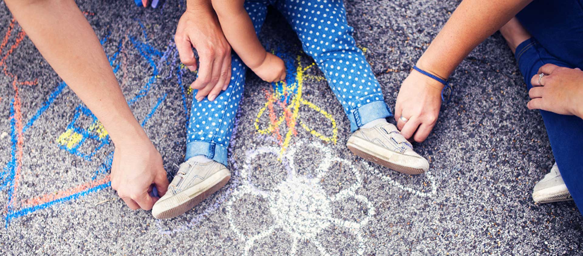 Two adults trace the legs of a preschooler using chalk on pavement.